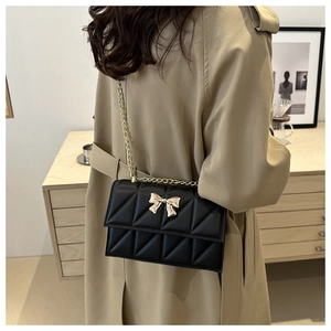 Quilted black crossbody bag with gold bow clasp shown worn over a beige trench coat