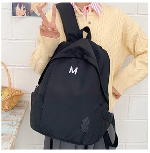 Detailed close-up of a black canvas backpack with white letter M print and brand tag