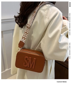 Brown camera bag with SM embossed detail and geometric wide fabric strap worn by a model