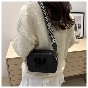 Close-up of black crossbody camera bag with wide geometric strap and golden hardware