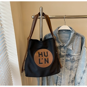 Detail of black canvas tote bag showing HU L·N logo patch and brown leather shoulder strap