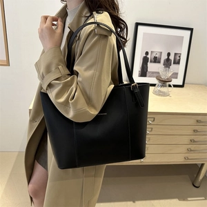 Model wearing black tote bag over beige coat showing strap detail and metal buckle