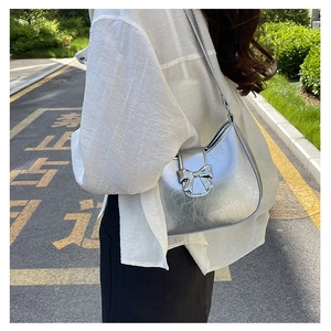Silver crescent shoulder bag with metallic bow detail shown in outdoor street setting
