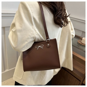 Brown leather tote bag with silver bow accent worn over the shoulder in a lifestyle setting