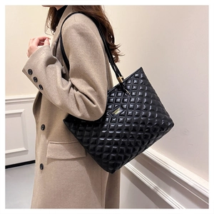 Detail of a black quilted tote bag with a gold metal plaque on camel coat background