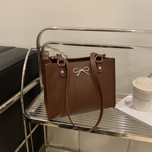 Brown leather tote bag with silver bow accent and shoulder straps on metal glass shelf
