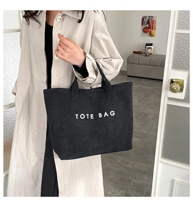 Detail einer schwarzen Wildleder-Tote-Bag mit weißem Aufdruck getragen zum beigen Trenchcoat