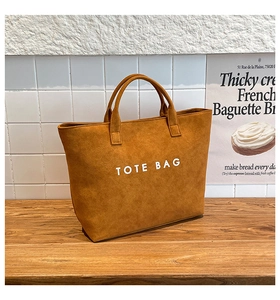 Klassische Shopper-Tasche aus braunem Wildleder mit weißem Aufdruck auf Holztisch