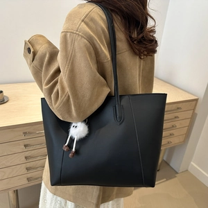 Minimalist black faux leather tote bag with plush charm shown on person in warm indoor setting