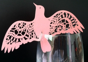 Pink laser-cut paper bird place card with floral lace patterns on a glass rim