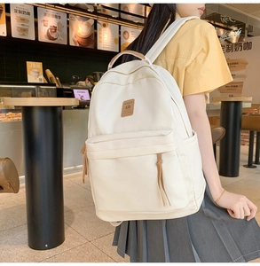 Cream canvas backpack detail featuring leather logo patch and zip puller in a cafe setting