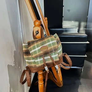 Green plaid woolen backpack with brown leather trim and drawstring pom-pom detail