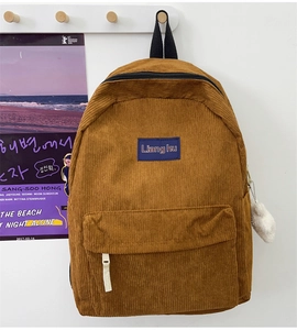Brown corduroy backpack detail featuring front pocket and white plush charm on a white wall