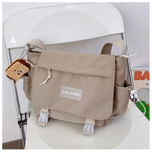 Détail du sac messager en nylon kaki avec logo et breloque biscuit sur une chaise blanche