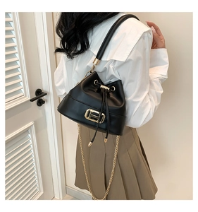 Detail of a black leather bucket bag with gold chain strap worn by a model in a room