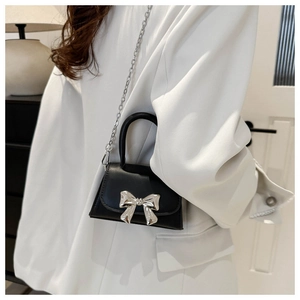 Mini black crossbody bag with silver bow detail worn by a model on a white blazer background