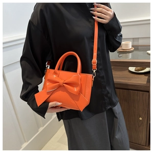 Detail of glossy orange handbag with large bow and crossbody strap on model