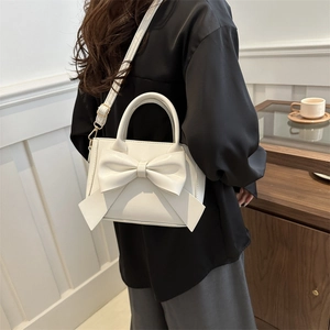 White trapezoidal crossbody bag with a large bow detail shown in a home setting
