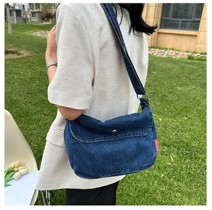 Detail of a large blue denim crossbody bag with metal buckle and red side label