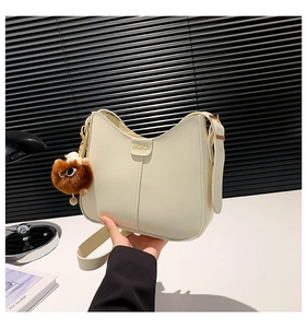 Detalle de bolso de hombro color crema con texto dorado y colgante de monstruo de peluche
