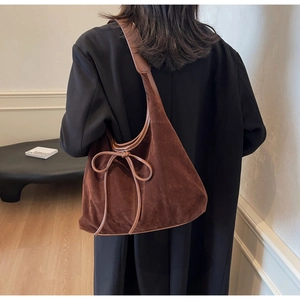 Close-up of a brown suede shoulder bag with a bow detail worn by a model in a cozy room