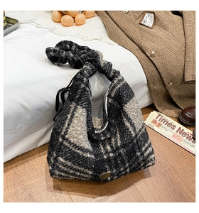 Detail der karierten Plüsch-Hobo-Tasche mit Knotengriff auf Holzboden und Zeitungsdeko