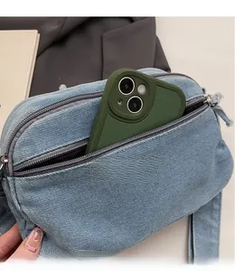 Detail of light blue denim crossbody bag showing multi-zipper storage with a smartphone
