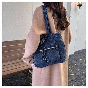 Detailed view of blue denim tote bag with front zip pockets and fringe tassels worn with coat