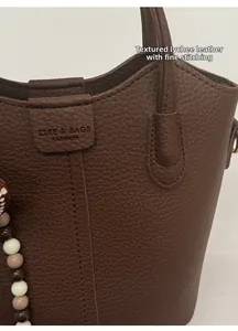 Detail einer braunen Eimertasche aus genarbtem Leder mit Kontrastnaht und Perlenanhänger