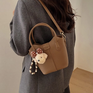 Dunkel khakifarbene Beuteltasche mit Teddybär-Anhänger an einem Model im grauen Anzug