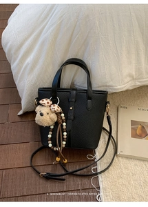 Detail of black lychee grain tote bag with plush bear charm on a wooden floor