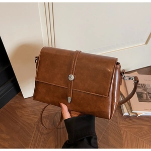 Detail of a brown leather messenger bag with silver round buckle in a cozy home setting