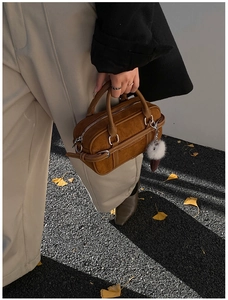Detailed view of a brown suede Boston bag with plush ball charm on an autumn street