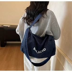 Close-up of indigo denim crossbody bag featuring white REPLACED text and wide shoulder strap