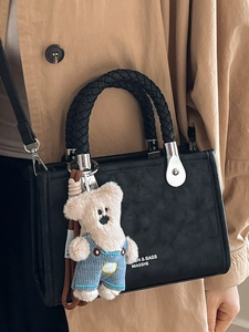 Black textured square tote bag with woven handle and plush bear charm on camel coat