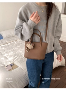 Brown leather bucket bag with teddy bear charm shown on a casual female model in a bedroom