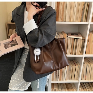 Detail view of large dark brown tote bag with white plush charm against a bookshelf
