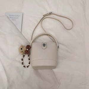 Close-up of cream bucket bag with metal buckle, shoulder strap and plush bear charm