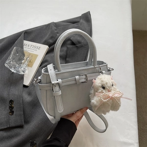 Detail of grey square crossbody bag with metal buckle and plush bunny charm on white fabric