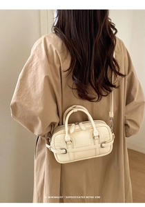 Sac Boston beige minimaliste porté en bandoulière sur un trench-coat dans un cadre chic