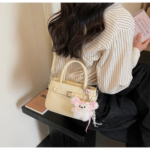 Detail of cream crossbody bag with leather buckle and plush doll charm in casual setting