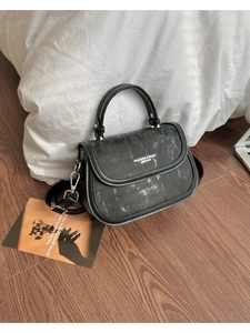 Détail du sac bandoulière en cuir noir avec boucle argentée sur fond de parquet