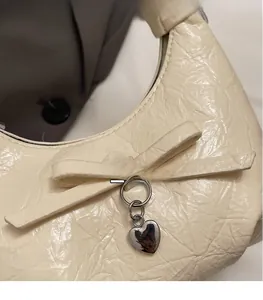 Détail d'un sac plissé crème avec un nœud en cuir et un pendentif en cœur argenté