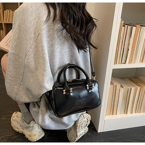 Detail of black glossy Boston bag with gold hardware and embossed logo on grey outfit