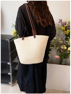 Detail of a beige woven straw tote bag with brown leather handles worn by a model