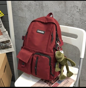 Detail of a dark red multi-pocket backpack on a chair with a green plush frog pendant