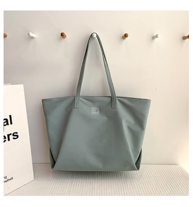 Detail of a sage green nylon tote bag featuring a white square logo on a minimalist wall