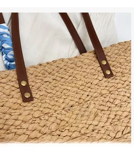 Detail of woven straw tote featuring brown leather straps, gold rivets, and a blue scarf