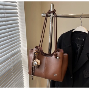 Detail of a brown tote bag featuring metal letter hardware and a plush pompom charm accent