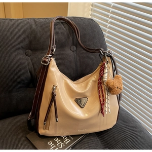 Beige Hobo-Tasche mit braunen Riemen und Plüschbär-Anhänger auf grauem Stoffsofa